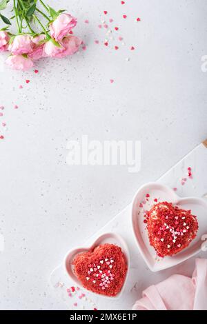 Zwei Kuchen Roter Samt in Form von Herzen auf weißem Teller, Rosenblumen und eine Tasse Kaffee auf rosafarbenem romantischem Hintergrund. Dessert-Idee für den Valentinstag, Stockfoto