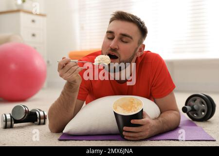 Fauler junger Mann, der Eis isst, anstatt zu Hause zu trainieren Stockfoto