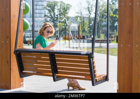 Porträt einer lächelnden Schülerin mit Sonnenbrille, die ein Smartphone in der Hand hält, auf einer Bank in einem Stadtpark sitzt. Ein glückliches Mädchen, das aus der Ferne lernt Stockfoto