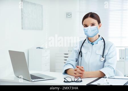 Junger Arzt trägt medizinische Maske am Schreibtisch im Büro Stockfoto