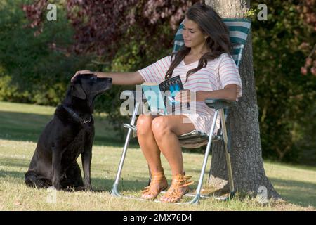 Junge Frau, die im Garten sitzt und ein Buch liest, mit schwarzem labrador Stockfoto