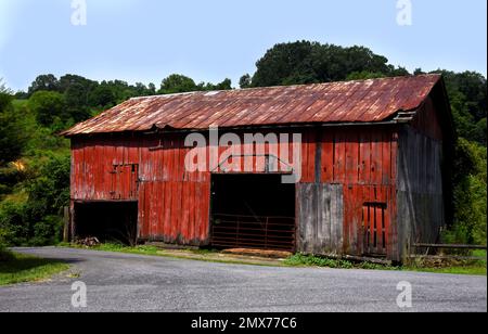 Rot, verblasst, verwittert, Holzscheune hat rostende und geknickte Blechdächer. Die Landstraße verläuft vor der Scheune. Stockfoto