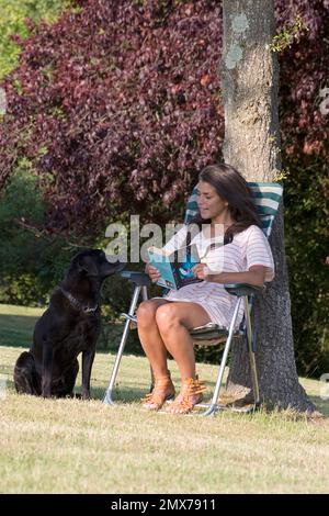 Junge Frau, die im Garten sitzt und ein Buch liest, mit schwarzem labrador Stockfoto