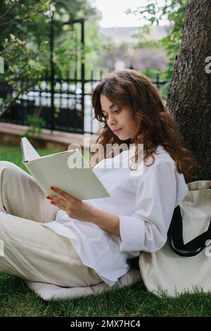 Bildmodell eines Magazins oder Buches. Das Mädchen entspannt sich auf dem Rasen im Hof des Cafés, liest ein Buch. Stockfoto