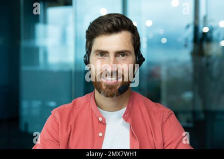 Nahaufnahme eines erwachsenen Mannes mit Bart und Headset für Videogespräche im Büro, Mitarbeiter des technischen Supports lächeln und in rotem Hemd auf die Kamera schauen, Online-Kundendienst und -Support. Stockfoto