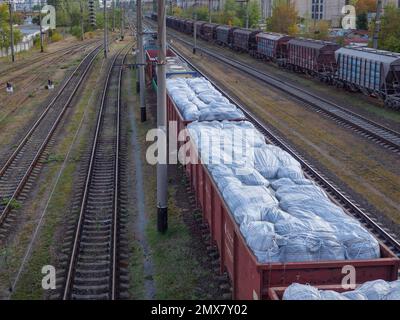 Ein Güterzug mit Güterwagen ohne Dächer, beladen mit riesigen weißen Säcken und wartet am Bahnhof in Kiew-Stadt, Ukraine. Stockfoto