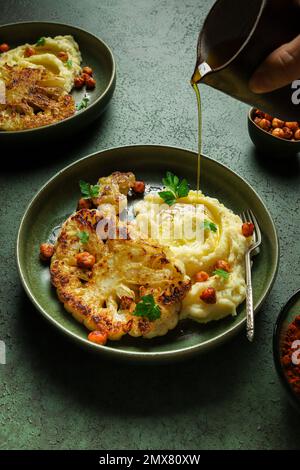 Körperteil eines nicht wiedererkennbaren Weibchens, das Olivenöl auf gebratenen Blumenkohl mit Kartoffelpüree und gewürzten Harissa-Kichererbsen mit frischem Pastinaken gießt Stockfoto