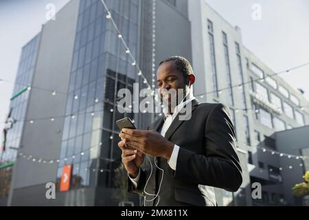 Foto von unten. Ein afroamerikanischer Geschäftsmann in einem Anzug steht vor einem Büro, trägt Kopfhörer und benutzt das Telefon. Hört zu, wählt, ruft an, redet, lächelt. Stockfoto