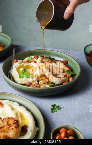 Körperteil eines nicht wiedererkennbaren Weibchens, das Olivenöl auf gebratenen Blumenkohl mit Kartoffelpüree und gewürzten Harissa-Kichererbsen mit frischem Pastinaken gießt Stockfoto