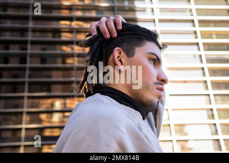 Kleiner Seitenwinkel von einem jungen Mann mit Dreadlocks, die wegschauen, während sie Haare berühren und vor einem modernen Gebäude stehen Stockfoto