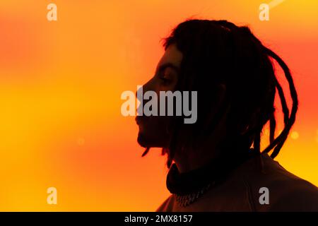 Seitenansicht eines pensiven Mannes in Dreadlocks, der gegen orangefarbenes Neonlicht steht und im Studio durchdacht wegschaut Stockfoto