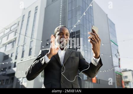 Foto von unten. Ein afroamerikanischer Geschäftsmann im Anzug steht vor einem Büro, trägt Kopfhörer und hält ein Telefon. Spricht bei einem Videoanruf, sagt Hallo, lächelt, schaut in die Kamera. Stockfoto