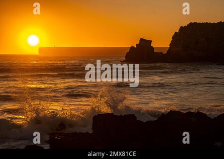 Malerischer Blick auf den hellen orangefarbenen Sonnenuntergang über dem Wellenwasser in der Nähe von Felsen und der Silhouette des Leuchtturms Sagres in Portugal Stockfoto