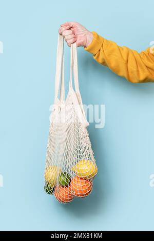 Seitenansicht einer verkürzten, nicht erkennbaren Seniorin im gelben Pullover mit Netztasche mit Früchten vor blauem Hintergrund Stockfoto