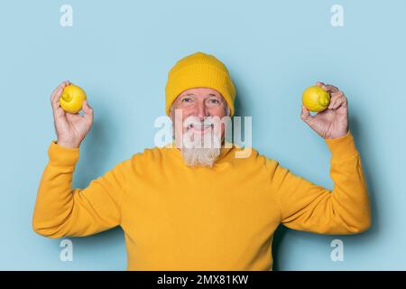 Porträt eines lächelnden Seniorenmannes mit Bart in gelbem Pullover und Hut mit frischen Zitronen, die vor blauem Hintergrund in die Kamera schauen Stockfoto