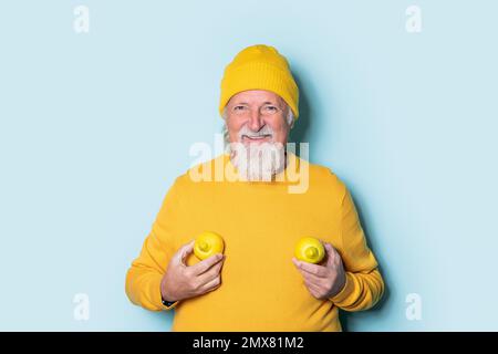 Porträt eines fröhlichen bärtigen älteren Mannes in gelbem Pullover und Hut mit zwei Zitronen auf der Brust, der vor blauem Hintergrund in die Kamera schaut Stockfoto