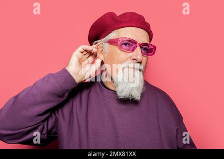 Porträt eines durchdachten männlichen Hipsters mit Bart in trendiger Sonnenbrille und Baskenmütze, der wegblickt und das Ohr auf pinkfarbenem Hintergrund berührt Stockfoto