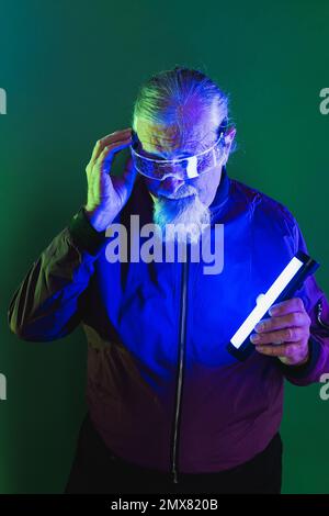 Überraschter männlicher Hipster mit Bart in Brille und lässiger Kleidung, die den Kopf berührt, während er im Studio vor dunklem Hintergrund mit Neonlicht steht Stockfoto