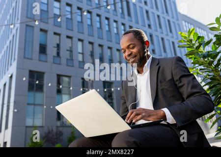 Foto von unten. Ein afroamerikanischer Mann im Anzug sitzt vor einem Büro auf einer Bank, auf einer Straße, trägt Kopfhörer und arbeitet online an einem Laptop, tippt und lächelt. Stockfoto