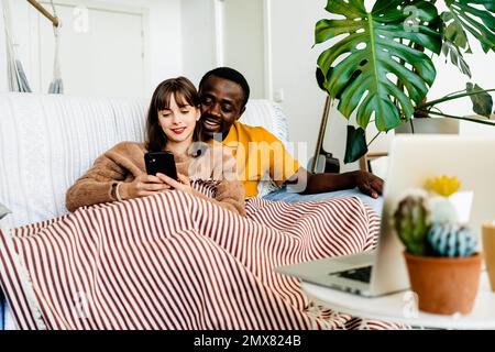 Glückliches junges, vielseitiges Paar in legerer Kleidung, das lächelt und im Smartphone surft, während es sich zu Hause auf dem Sofa entspannt Stockfoto