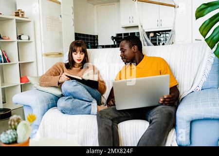 Glückliches multiethnisches Paar in lässiger Kleidung, das auf dem Sofa sitzt, ein Buch liest und im Wohnzimmer zu Hause ein Notebook benutzt Stockfoto