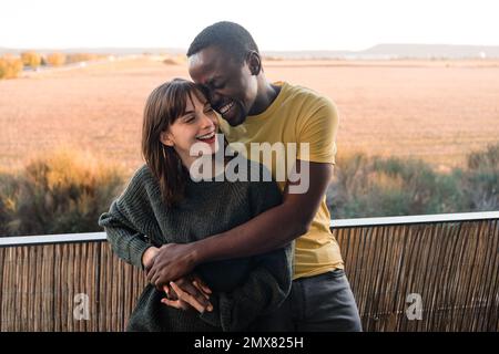 Ein entzücktes, multiethnisches Paar in legerer Kleidung, das lächelt und sich umarmt, während es auf dem Balkon des Hauses steht und sich gegen den Sonnenuntergang lehnt Stockfoto