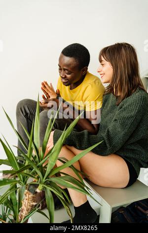 Ein fröhliches, multiethnisches Paar in legerer Kleidung, das lächelt und Händchen hält, während es auf Treppen neben einer Topfpflanze an einer weißen Wand sitzt Stockfoto