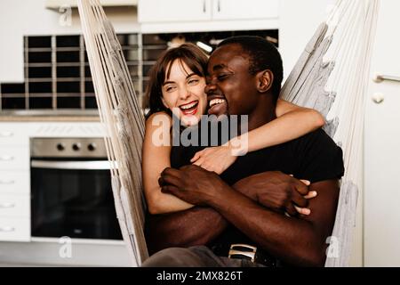 Fröhliches, multiethnisches Paar in lässiger Kleidung, das sich umarmt und lacht, während es am Wochenende auf dem Schaukelstuhl zusammen in der Küche chillt Stockfoto