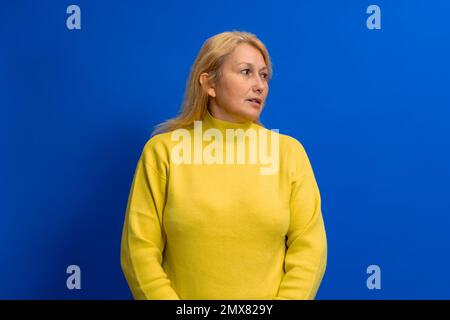Wunderschöne Frau in ihren 40s Jahren, die einen gelben Pullover trägt, auf blauem Hintergrund steht und auf die Seite schaut, mit einem ernsthaften Gesicht. Stockfoto