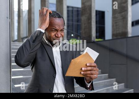 Ein aufgebrachter afroamerikanischer Mann stand in der Nähe des Gerichtsgebäudes und hielt einen Umschlag mit einem Brief in der Hand. Er hält seinen Kopf, erhielt Scheidungsdokumente, finanzielle Vereinbarung, Unterhalt, Klage. Stockfoto