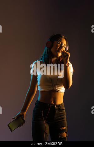 Selbstbewusste junge Frau in trendigen Outfits hört Musik über Kopfhörer und singt vor dunklem Hintergrund Stockfoto