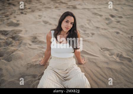 Von oben positiv junge hispanische Touristinnen mit langen dunklen Haaren oben und Rock, die auf dem Sandstrand mit gekreuzten Beinen in die Kamera schauen Stockfoto