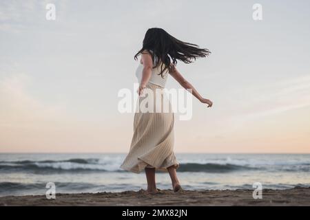 Rückblick auf eine sorglose junge, nicht erkennbare barfüßige Frau mit langen dunklen Haaren in Sommerkleidung, die auf einem Sandstrand in der Nähe von schaumigen Tänzen tanzt Stockfoto