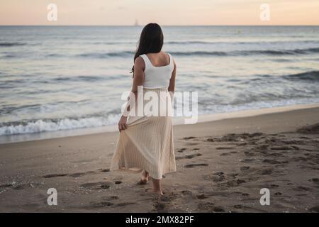 Rückblick auf eine sorglose junge, nicht erkennbare barfüßige Frau mit langen dunklen Haaren in Sommerkleidung, die am Sandstrand neben einem schäumenden Schaum stand Stockfoto