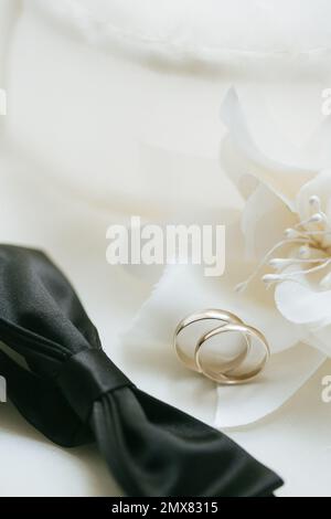 Von oben elegante Eheringe neben schwarzer Fliege und weißer zarter Blume auf weißer Oberfläche Stockfoto