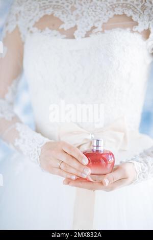 Nicht wiedererkennbare Braut in weißem Hochzeitskleid mit Parfümflasche in den Händen auf verschwommenem Hintergrund Stockfoto