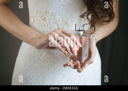 Nicht wiedererkennbare Braut in weißem Hochzeitskleid mit Parfümflasche in den Händen auf verschwommenem Hintergrund Stockfoto