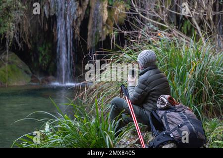 Rücksichtsvoller älterer männlicher Wanderer in warmer Kleidung und Rucksack, der auf dem See mit Thermoskannen sitzt und wegschaut, während er die Natur genießt Stockfoto