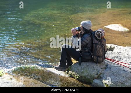 Der ganze Körper einer anonymen weiblichen Reisenden in Oberbekleidung und Hut mit Rucksack sitzt auf einem Stein in der Nähe des Sees mit Wanderstöcken, während sie ein heißes Getränk trinken Stockfoto