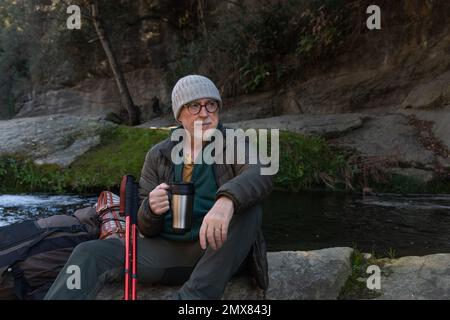 Rücksichtsvoller älterer männlicher Wanderer in warmer Kleidung und Rucksack, der auf dem See mit Thermoskannen sitzt und wegschaut, während er die Natur genießt Stockfoto