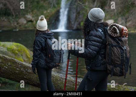 Nicht wiedererkennbare Touristen in warmer Kleidung mit Rucksäcken und Wanderstöcken stehen am Ufer des Sees in der Nähe des Wasserfalls während der gemeinsamen Reise Stockfoto