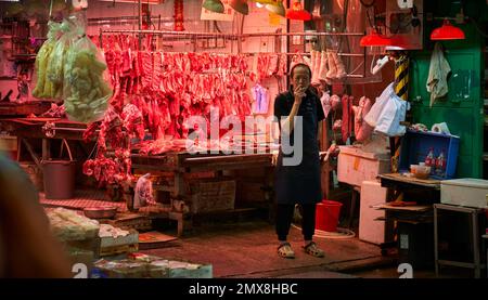 Ein Metzger, der eine Zigarette raucht, während er vor seinem Stand auf einem Straßenmarkt in Hongkong steht. Stockfoto