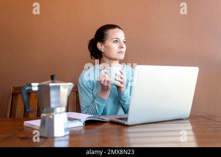 Frau sitzt in ihrem Pyjama, arbeitet an ihrem Laptop und trinkt eine Tasse Kaffee. Stockfoto