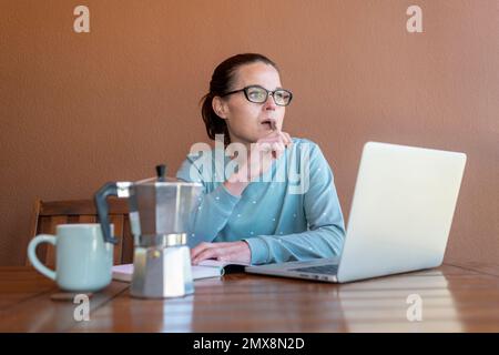 Frau, die in ihrem Pyjama sitzt, an ihrem Laptop arbeitet und wegschaut und denkt. Stockfoto