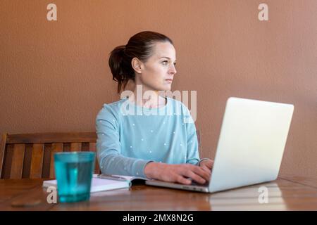 Frau, die in ihrem Pyjama sitzt, an ihrem Laptop arbeitet und wegschaut und denkt. Stockfoto