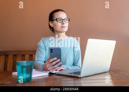 Eine Frau, die in ihrem Pyjama sitzt und an ihrem Laptop und Handy arbeitet und wegschaut und denkt. Stockfoto