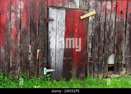 Die alte Scheunentür ist geschlossen und wird mit einem Holzschloss und rostigen Scharnieren gehalten. Das Hintergrundbild zeigt gerissene, verblasste und verwitterte Platten. Stockfoto
