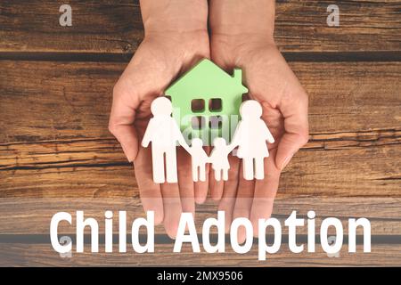 Frau hält Figuren von Familie und grünem Haus in den Händen auf Holzhintergrund, Draufsicht. Adoptionskonzept Stockfoto