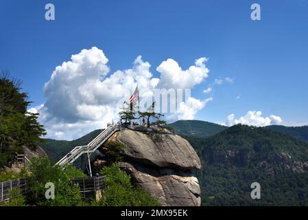 Vom Gipfel des Chimney Rock im Chimney Rock State Park haben Besucher einen atemberaubenden Blick. Die amerikanische Flagge steht oben. Wolken umrahmen die Flagge und Smokey Mountai Stockfoto