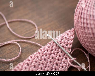 Rosafarbenes Baumwollgestrickmaterial, Häkelhaken mit Metallgriff und Garn auf dem Holztisch, Nahaufnahme, selektiver Fokus. Häkelpraxis Stockfoto